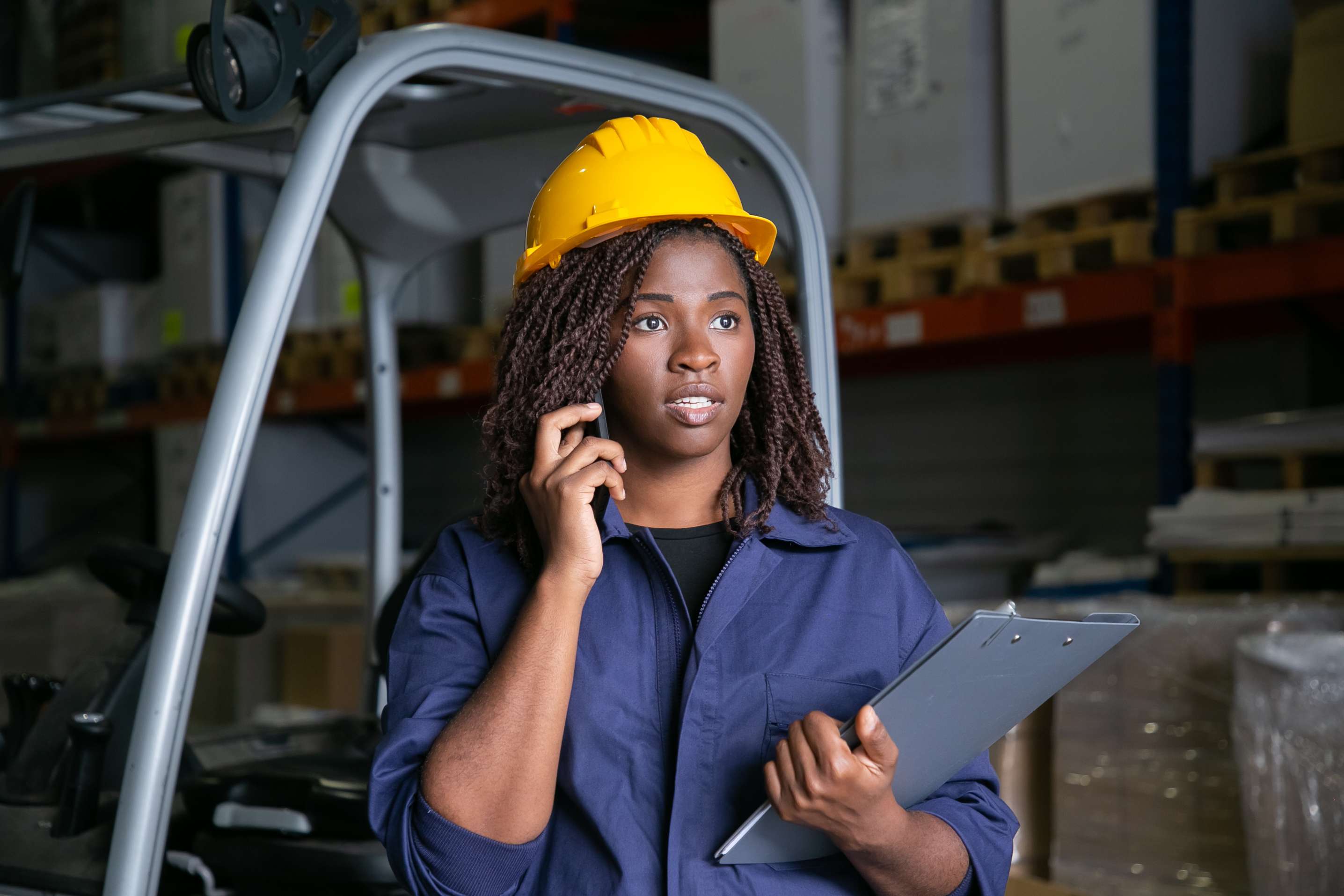focused-black-warehouse-worker-yellow-hardhat-standing-near-forklift-talking-cell-shelves-with-goods-background-medium-shot-labor-communication-concept (1)